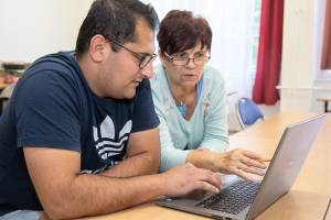 Ein Mann mittleren Alters mit Brille und dunkelblauem T-Shirt sitzt an einem Tisch und arbeitet konzentriert an einem Laptop. Neben ihm sitzt eine ältere Frau mit kurz geschnittenem, rotbraunem Haar und Lesebrille, die ihm etwas auf dem Bildschirm zeigt. Sie trägt eine hellblaue Bluse. Beide wirken fokussiert und im Gespräch vertieft. Der Raum ist hell, im Hintergrund stehen weitere Stühle und ein Fenster mit roten Vorhängen.