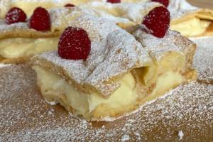 Foto eines Stückes Karpatka-Kuchen mit Himbeeren. Die hügelige Oberfläche ist namensgebend für den Kuchen. Die mit Puderzucker bestäubten Wellen erinnern an Hügellandschaften in den Karpaten.