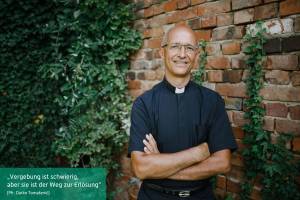 Das Bild zeigt einen Priester, der vor einer Ziegelmauer steht, die teilweise mit Efeu bewachsen ist. Er lächelt, trägt eine Brille und die traditionelle schwarze Priesterkleidung mit einem weißen Kollar. Seine Arme sind verschränkt, und er wirkt entspannt. Die Umgebung hat eine friedliche und natürliche Atmosphäre, mit Grünpflanzen, die die Wand emporranken. Unten links im Bild befindet sich ein Zitat: „Vergebung ist schwierig, aber sie ist der Weg zur Erlösung“ (Pfr. Darko Tomašević)