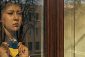 Das Bild zeigt ein junges Mädchen mit ernstem, nachdenklichem Gesichtsausdruck, das durch ein Fenster schaut. Sie hält ein Herz aus Papier in den Farben der ukrainischen Flagge – oben blau, unten gelb – vor ihrer Brust. Die Reflexion im Fenster zeigt kahle Bäume und ein Gebäude im Hintergrund.